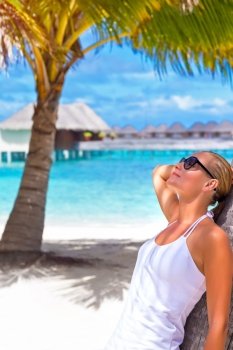Cute blond girl leaned on palm tree and looking on the sun through sunglasses, summer time vacation, leisure concept