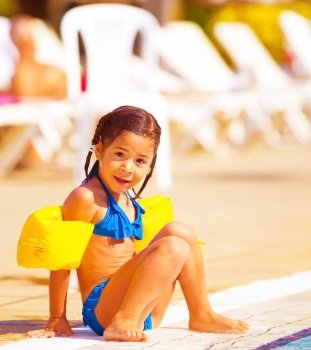 Cute little girl sitting near pool, active happy childhood, summer holidays, having fun outdoors, wearing swimwear, joy and pleasure concept