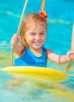 Cute happy baby girl enjoying summer holiday in the pool, having fun on water swing, red frangipani flower in hair, yellow sun print on cheek, summer vacation concept