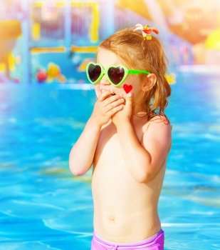 Closeup on sweet baby girl having fun in swimming pool, adorable child with red heart paint print on cheek wearing cute sunglasses, summer holidays in aquapark