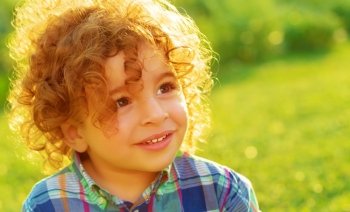 Closeup portrait of cute baby boy with curly hair on green field, having fun outdoors, enjoying summer vacation, happy childhood concept