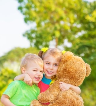 Two happy children enjoying big brown soft bear outdoors, brother and sister hugging, having fun on spring time, love and happiness concept
