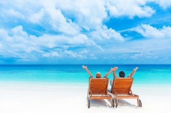 Two happy people relaxing on the beach, sitting on comfortable sunbed with raised up hands for joy, rear view, summer holidays concept   
