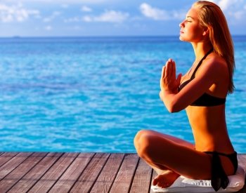 Woman meditating on the beach, doing exercise on wooden deck over blue sea, summer vacation, yoga and relaxation concept
