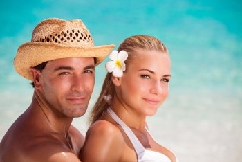 Closeup portrait of beautiful young lovers on the beach, handsome guy wearing straw hat hugging his nice girlfriend with flower in hair, romantic summer vacation