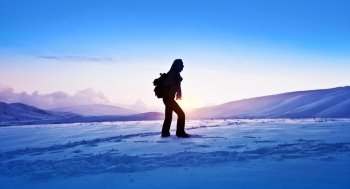 Woman traveler hiking in winter mountains, trekking in wintertime cold snowy weather, girl silhouette over natural blue sky with bright sunset and beautiful landscape, freedom concept