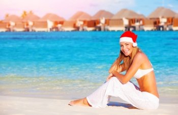 Christmas vacation on Maldives, pretty woman sitting on the beach, wearing red Santa hat, side view, luxury wintertime holidays concept