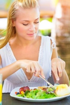 Portrait of attractive woman eating fresh salad and meat using knife and fork, having lunch in outdoors restaurant, healthy lifestyle concept