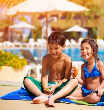 Two happy kids eating croissant near pool, having breakfast on the beach, active summer holidays, brother and sister enjoying sunny day, happiness concept