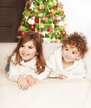 Photo of adorable kids, boy and girl laying down on the sofa and laughing, family at home celebrate Christmas, little babies playing indoor, infant Xmas party, decorated Christmas tree for children