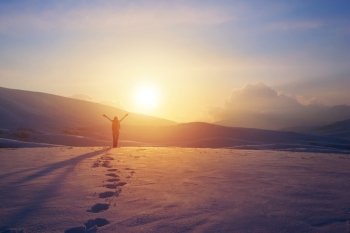 Happy joyful woman having fun outdoors in winter, standing on the mountains with raised up hands on the mountain covered with snow, enjoying beautiful sunset