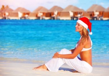 Beautiful young woman celebrating Christmas holiday on Maldive island, wearing red Santa hat, sitting on the beach, luxury resort