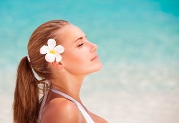 Side view portrait of beautiful calm female with frangipani flower in hair meditating on the beach, enjoying day spa, summer vacation and relaxation