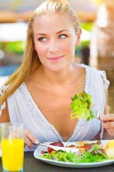 Portrait of attractive woman eating fresh green salad using knife and fork, having lunch in outdoors restaurant, healthy lifestyle concept