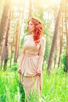 Dreamy girl standing in a forest on sunny spring day, wearing long elegant dress and flower wreath on curly red hair, fashion look