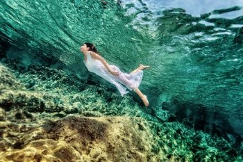 Woman swimming near rock in transparent blue sea, wearing white dress, relaxation in refreshing water, summer enjoyment concept