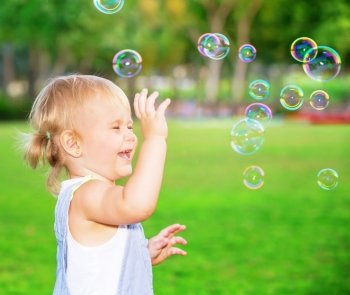 Happy child having fun in the park, cute blond baby girl playing with soap bubbles on the yard, joyful little kid enjoying outdoors game