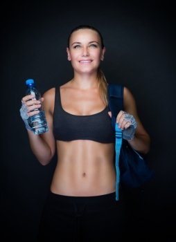 Sportive woman with backpack and bottle of water in hand over dark background, beautiful sportive trainer, active healthy lifestyle