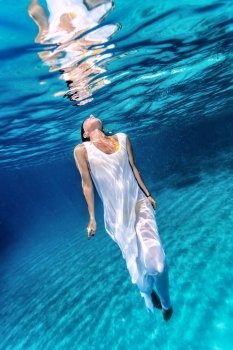 Beautiful female wearing white dress underwater, active lifestyle, diving in clear transparent water of sea, summer vacation concept