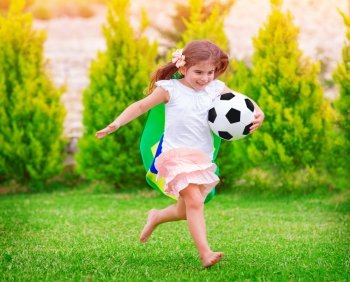 Little active football fan running on the green grass field with ball, young supporter of Brazilian football team with national flag 