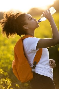 Outdoor portrait of beautiful happy mixed race African American girl teenager female young woman drinking water from a bottle in a field of yellow flowers at sunset in golden evening sunshine