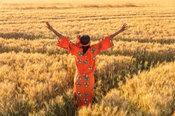 African woman in traditional clothes standing arms raised in a field of barley or wheat crops at sunset or sunrise