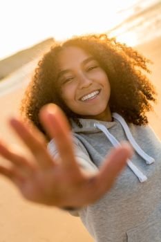 Outdoor portrait of beautiful happy mixed race African American girl teenager female young woman on a beach reaching out to camera, smiling and laughing with perfect teeth in golden sunset evening sunshine
