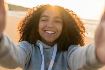 Outdoor portrait of beautiful happy mixed race African American girl teenager female young woman on a beach taking a selfie photograph smiling laughing with perfect teeth in golden sunset evening sunshine
