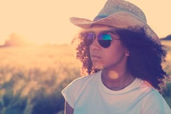 Instagram filter style photo of beautiful happy mixed race African American female girl teenager young woman wearing reflective aviator sunglasses and cowboy hat in a cornfield at golden sunset or sunrise