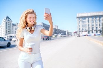 Beautiful young woman with music headphones and a take away coffee cup, taking picture of herself, selfie against urban city background.