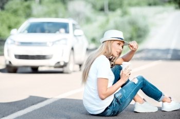 Beautiful young woman wearing hat and music headphones around her neck, drinking coffee from a takeaway coffee cup and sitting on a separating strip on the road.