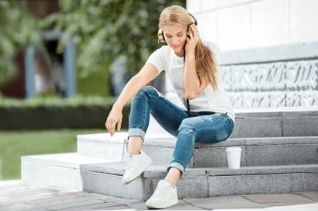 Happy young woman listening to the music in vintage music headphones and sitting on stairs with a take away coffee cup against urban city background.