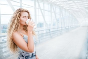 Beautiful young woman with long curly hair, holding a take away coffee cup and standing on the bridge against urban background.