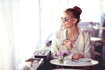 Photo of a beautiful happy young woman with smartphone and sunglasses, drinking hot tea or coffee on a cafe patio and looking out the window.