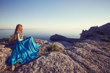 Young beautiful woman in blue dress looking to mountains sea. Girl on the nature on blue sky background. Fashion photo