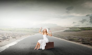 She is traveling light. Woman in white long dress and hat sitting on her luggage on asphalt  road