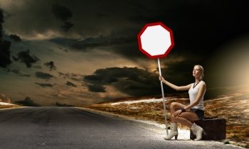 Girl with roadsign. Young woman sitting on suitcase and holding roadsign