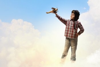 Image of little boy in pilots helmet playing with toy airplane against clouds background