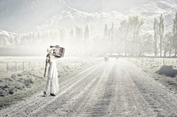 She is traveling light. Woman with suitcase in white long dress and hat on countryside road