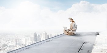 Writer woman working in isolation. Woman in dress and hat sitting on airplane wing and working on laptop