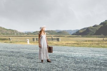 She is traveling light. Woman with suitcase in white long dress and hat on asphalt  road