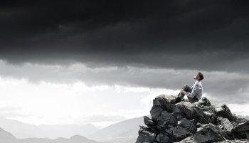 Isolated to find inspiration. Young smiling businessman sitting alone on rock top 