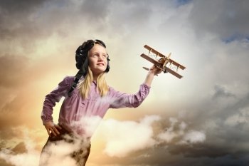 Little girl in pilot’s hat. Image of little girl in pilots helmet playing with toy airplane against clouds background