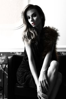 fashion portrait of beautiful woman with blue elegant shiny dress and fur on shoulders. Sitting on sofa in elegant ambient and looking in camera ,  black and white image with high contrast