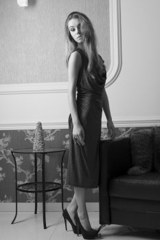 sophisticated charming woman in elegant interior ambient with long hair, make-up and shiny blue dress posing near sofa and looking in camera with haughty expression BW image