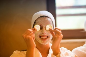 Beautiful young woman is getting facial clay mask at spa, lying with cucumbers on eyes