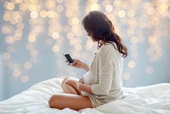 pregnancy, motherhood, technology, people and expectation concept - pregnant woman with smartphone in bed over holidays lights background
