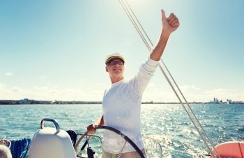 sailing, age, tourism, travel and people concept - happy senior man in captain hat on steering wheel and showing thumbs up sail boat or yacht floating in sea