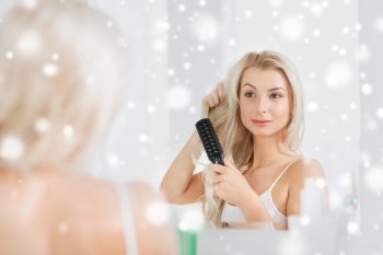 beauty, grooming, hair care and people concept - smiling young woman looking to mirror and brushing hair with comb at home bathroom over snow