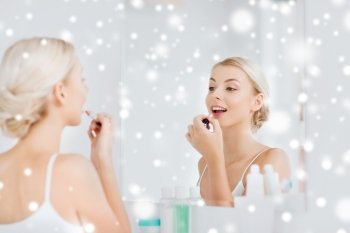 beauty, make up, cosmetics, morning and people concept - smiling young woman coloring her lips with lipstick  looking to mirror at home bathroom over snow